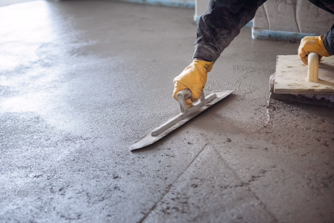 Worker using a metal trowel to level fresh screed on a concrete floor, smoothing the surface for further flooring installation.