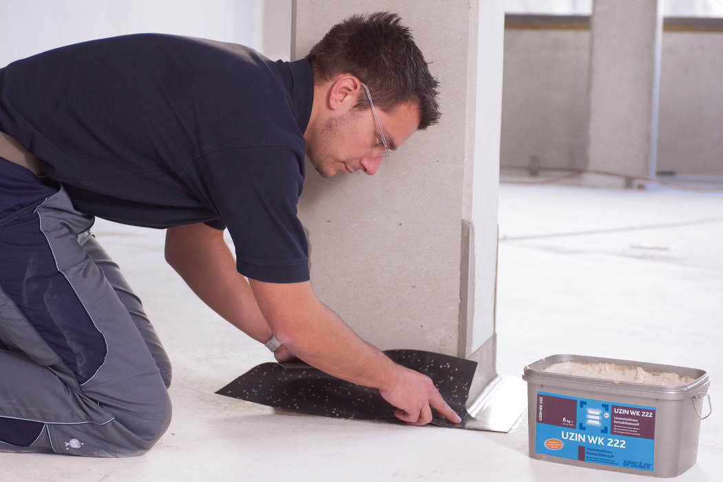 A construction worker wearing safety glasses and work clothes kneels inside a clean, unfinished interior and presses a black adhesive membrane or underlayment at the base of a light-colored wall or column. He smooths the material with his hands along the floor edge. Nearby on the floor is a plastic container labeled 'UZIN WK 222' containing adhesive. The scene shows a concrete or screed floor with minimal background details.