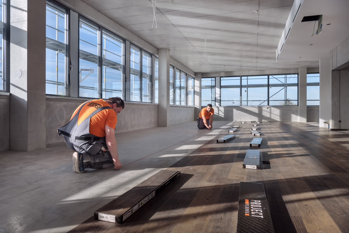 Two installers laying wood-look LVT flooring in a spacious, light-filled room with large windows and prepared subfloor surfaces.