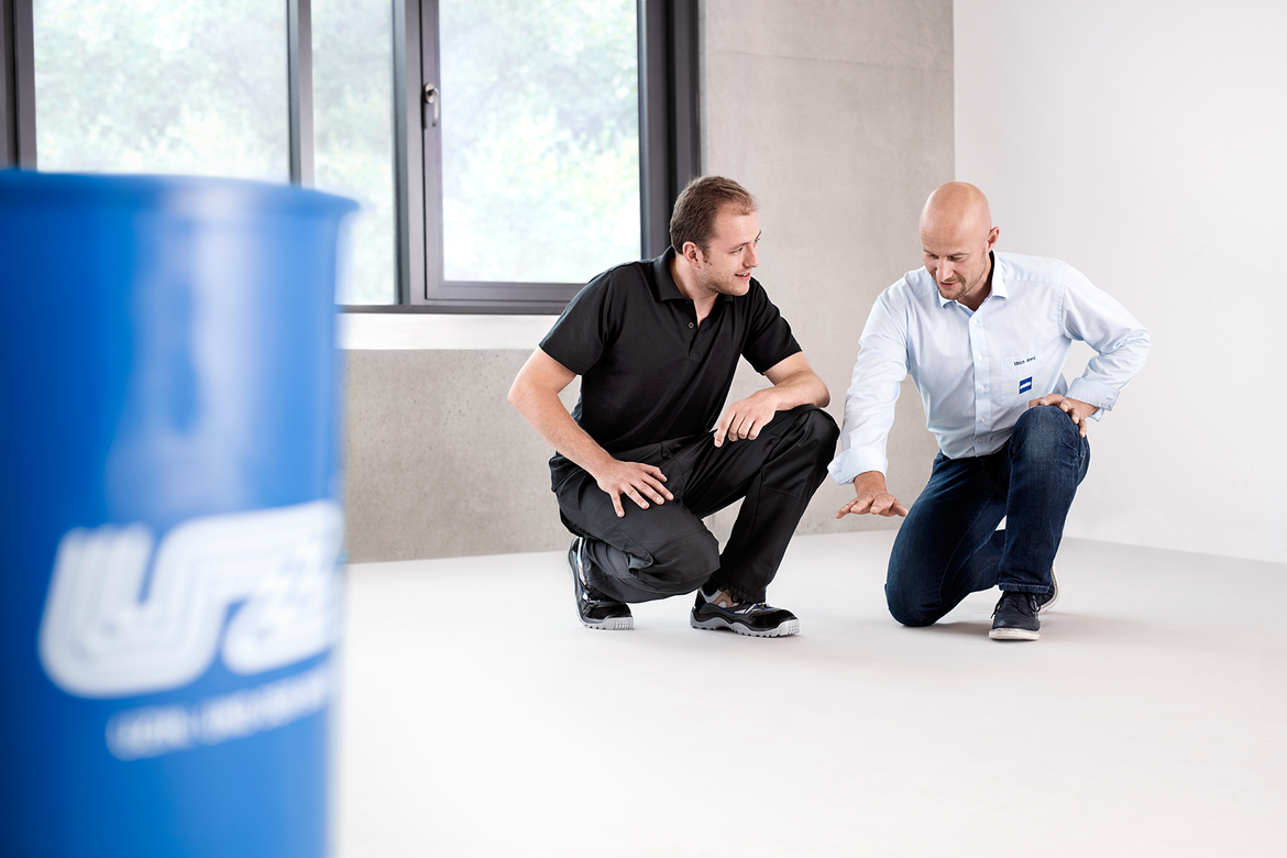 Two men kneel on a light-colored floor; one in a black shirt, the other in a light blue shirt, pointing at the floor. A blue barrel with white markings is visible on the left side of the image.