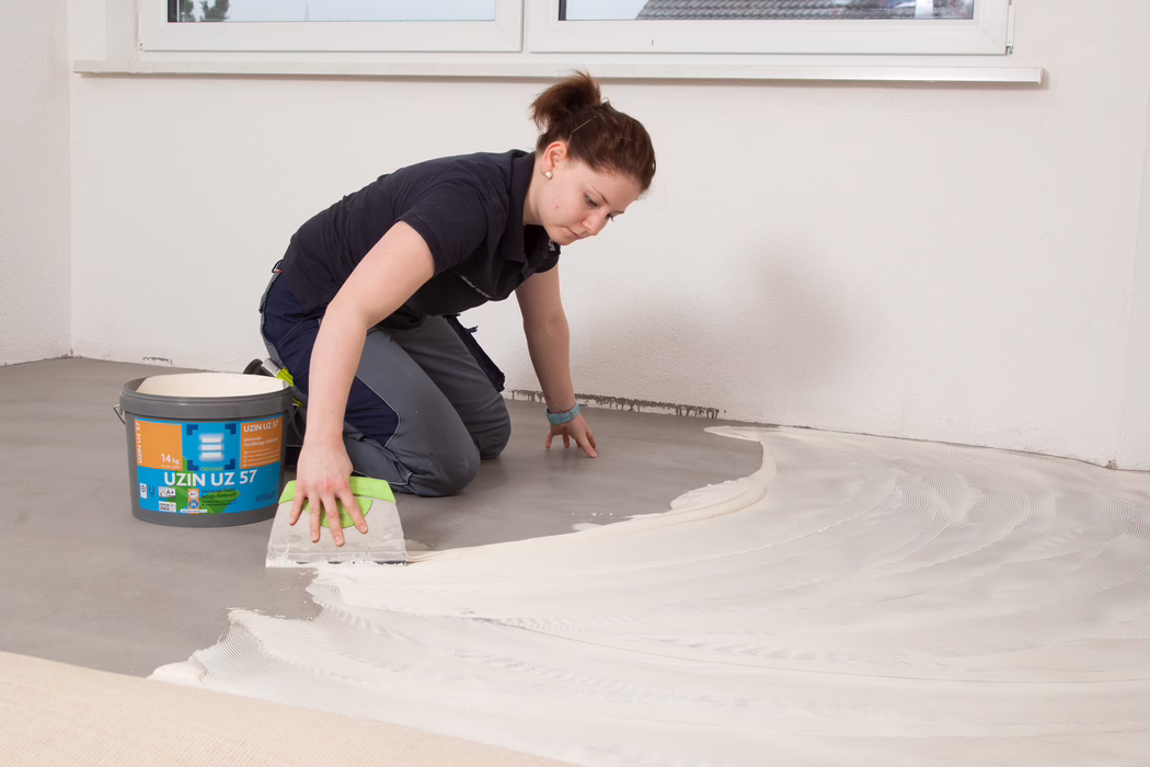 UZIN_Application Image_UZ57_1_6182_2018-11 A person is kneeling on a bare concrete floor inside an unfurnished room, spreading a light-coloured adhesive or levelling compound with a wide notched trowel. A bucket labelled "UZIN UZ 57" stands nearby; the material is being applied in sweeping arcs toward the wall under a window. The worker wears dark work clothes, has their hair tied back, and focuses on creating an even layer for a subsequent floor covering.