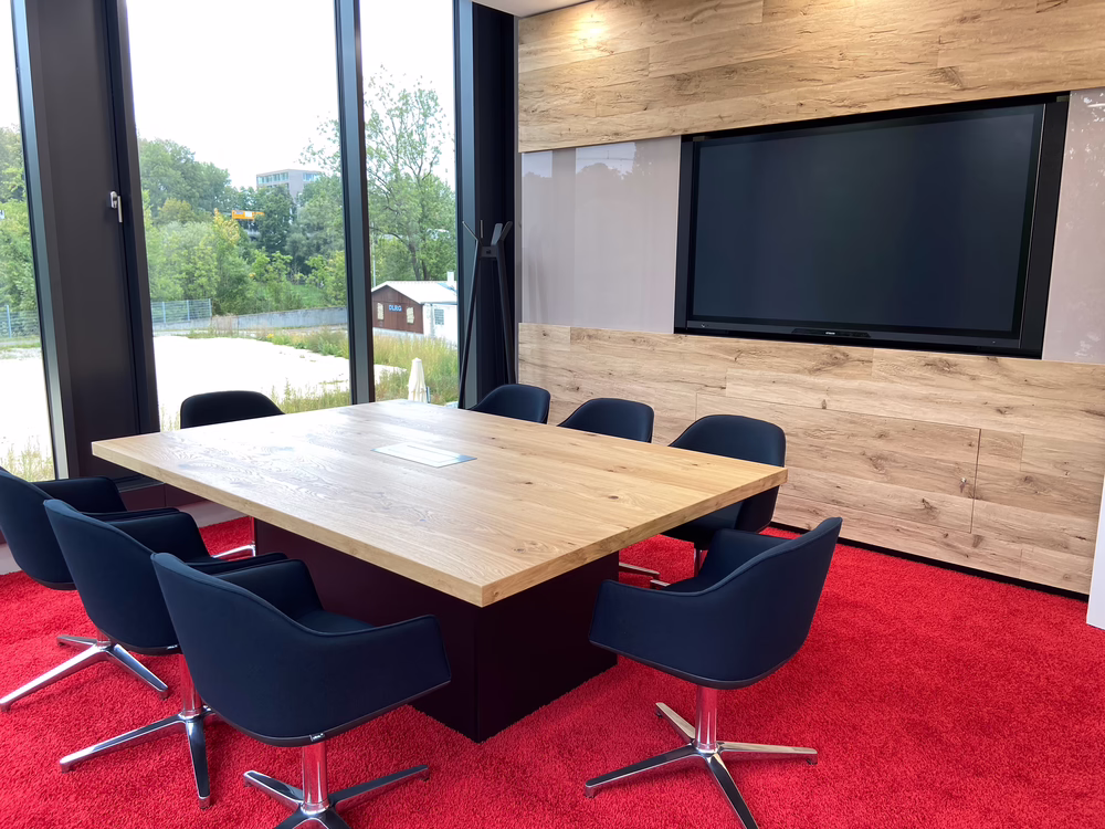 Meeting room with red carpet flooring, wooden conference table, blue upholstered chairs, and a large wall-mounted screen.